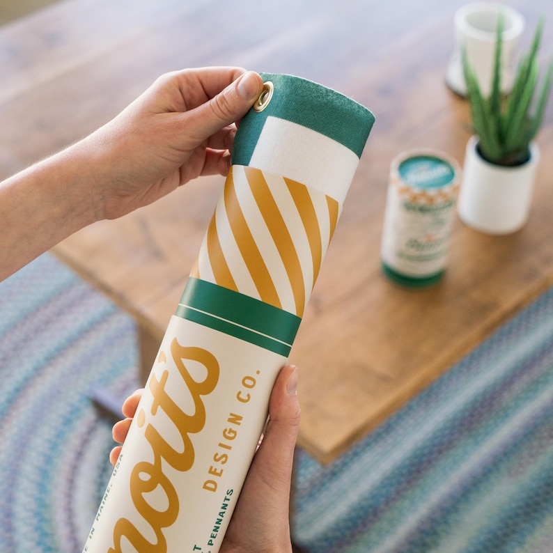 A person's hands holding a cylindrical object with a striped pattern, which appears to be a cylindrical container or tube. The background includes a wooden surface and some plants, creating a natural and indoor setting.
