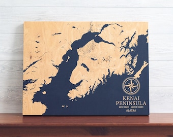 May include: A wooden map of the Kenai Peninsula in Alaska, with a blue water feature and a compass rose. The text "Kenai Peninsula, West Coast - United States, Alaska" is printed below the compass rose.
