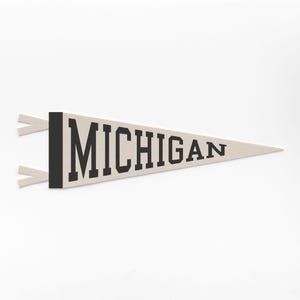 Op de afbeelding: Een beige wimpel met het woord "MICHIGAN" in vette zwarte letters. De wimpel heeft een zwarte verticale streep aan de linkerkant en twee beige linten. De wimpel is driehoekig van vorm en wordt weergegeven tegen een witte achtergrond.