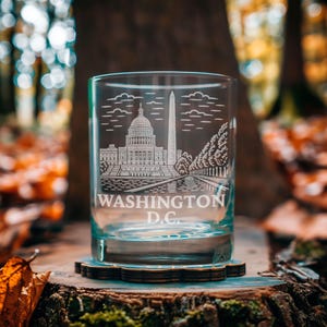 May include: A clear glass tumbler with an etched design of Washington D.C. landmarks, including the Capitol Building and Washington Monument. The text "WASHINGTON D.C." is etched below the image. The glass sits on a wooden coaster.