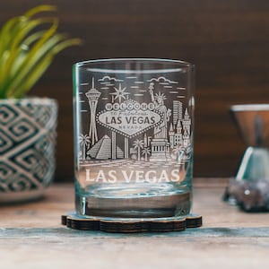 Może przedstawiać: Przezroczysty szklany kubek z wygrawerowanym wzorem Las Vegas, zawierającym napisy "Welcome to Fabulous Las Vegas Nevada" i kultowe zabytki. Szklanka stoi na dekoracyjnej podstawce.