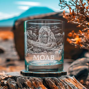 May include: A clear glass tumbler with a detailed etched design featuring a landscape with a road, a jeep, a bicycle, and the word "MOAB". The design is in white against the clear glass. The glass sits on a wooden coaster.
