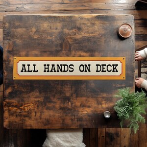 Puede incluir: Un letrero rectangular con el texto "ALL HANDS ON DECK" en letras negras. El letrero tiene un fondo crema con un borde amarillo y está montado sobre una superficie de madera oscura. El letrero es un objeto decorativo.