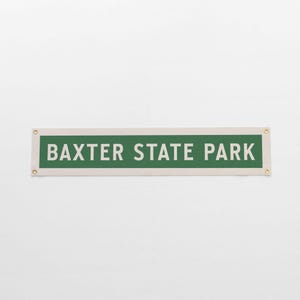 May include: A rectangular sign with the text "BAXTER STATE PARK" in white letters on a green background. The sign is bordered by a cream-colored frame and has brass grommets in each corner.
