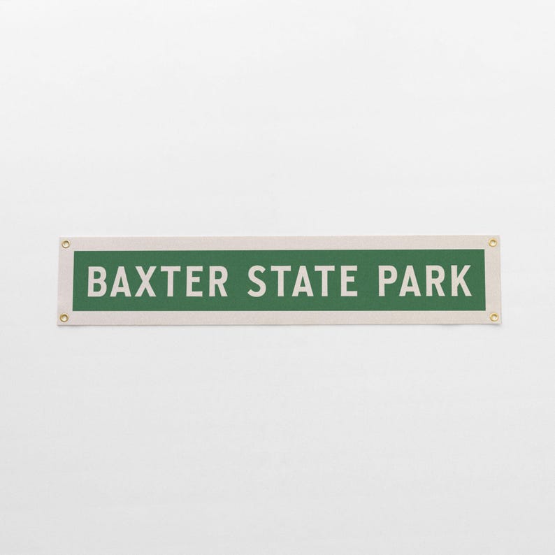 May include: A rectangular sign with the text "BAXTER STATE PARK" in white letters on a green background. The sign is bordered by a cream-colored frame and has brass grommets in each corner.
