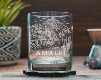 Op de afbeelding: Een helder glazen tumbler met een gedetailleerd geëtst ontwerp van de Amalfikust, inclusief gebouwen, bergen, palmbomen en een boot. De tekst "AMALFI COAST" is eronder geëtst. Het glas staat op een houten onderzetter.