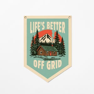 May include: A cream-colored fabric wall banner with a pointed bottom edge and metal grommets. The graphic features a log cabin, pine trees, and a mountain against a red sun, with the text LIFE'S BETTER OFF GRID in a bold, retro-style font.