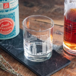 May include: A clear glass tumbler with a white etched design of the Pittsburgh skyline and the text "PITTSBURGH PENNSYLVANIA". The tumbler is sitting on a black slate coaster.
