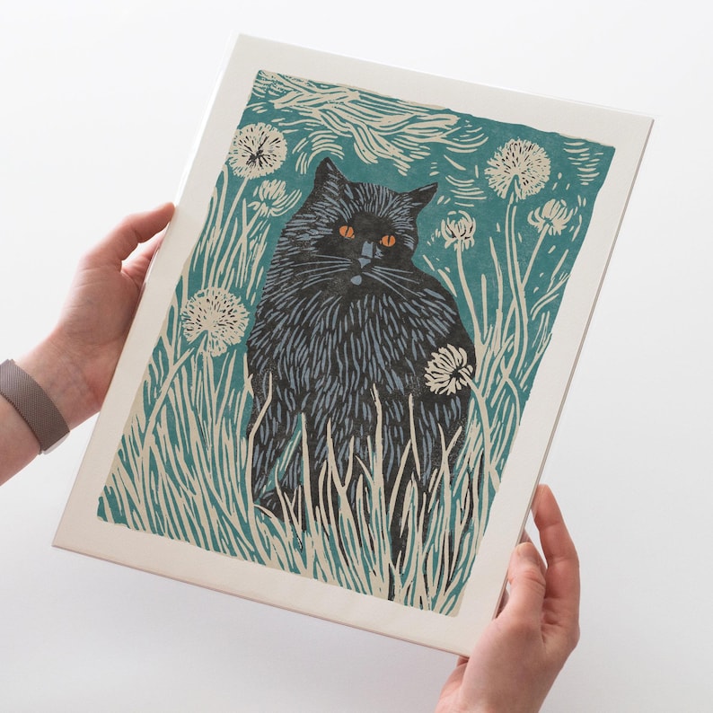 May include: A framed art print featuring a black cat with orange eyes, set in a field of tall grass and dandelions. The artwork uses a woodcut style with teal, white, and black colors. The cat is the central focus of the print.