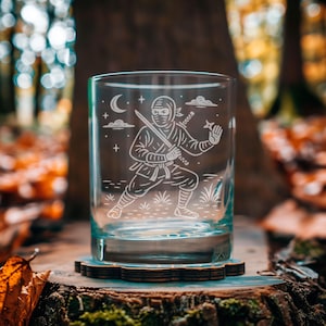 May include: A clear glass tumbler with a ninja design etched on the front. The ninja is depicted with a sword and throwing star, with a moon and stars in the background. The glass sits on a wooden coaster.