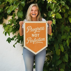 Può includere: Un banner arancione con le parole "Progress NOT Perfection" in corsivo bianco. Lo striscione è tenuto davanti a uno sfondo di foglie verdi. Lo striscione ha un bordo color crema ed è tenuto da due piccole puntine dorate.