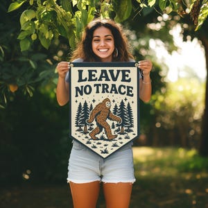 May include: A rectangular banner with the text "LEAVE NO TRACE" above an illustration of Bigfoot walking through a forest. The banner is cream-colored with a navy blue border and gold grommets. The Bigfoot is brown, and the trees are dark green.