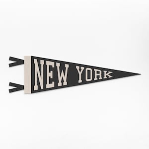 May include: A black pennant with the text "NEW YORK" in white block letters. The pennant has a triangular shape and is made of felt. Two black fabric strips are attached to the left side.