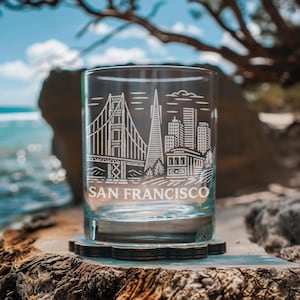 Puede incluir: Un vaso de vidrio transparente con un diseño grabado de los monumentos de San Francisco, incluyendo el Golden Gate Bridge y la Pirámide Transamerica. Las palabras "SAN FRANCISCO" están grabadas debajo. El vaso está sobre un posavasos de madera.