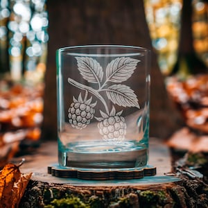 May include: A clear glass tumbler with a detailed etched design of a raspberry branch with leaves and two berries. The glass sits on a wooden coaster, with a blurred forest background.