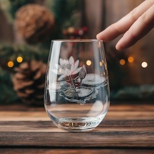 May include: A clear glass wine glass with an etched design of a white water lily with green leaves. The glass is sitting on a wooden table.