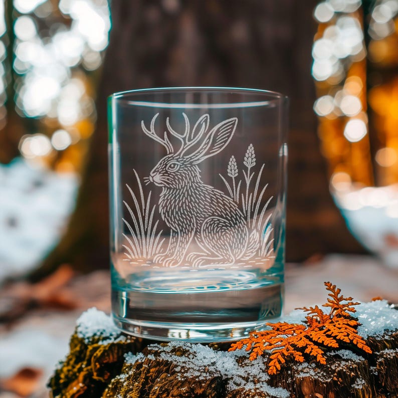 May include: A clear glass tumbler with a detailed etched design of a jackalope, a mythical creature with the body of a rabbit and antlers. The design includes tall grass and wheat. The glass is set on a mossy, snow-covered surface.