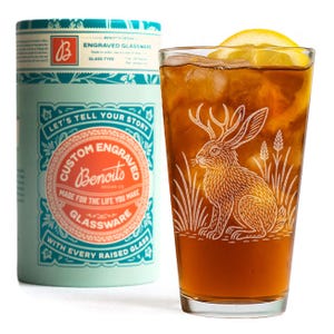 May include: A tall glass of iced tea with a lemon wedge, next to a cylindrical container. The glass is etched with a rabbit design. The container is light blue with the text "Custom Engraved Glassware".