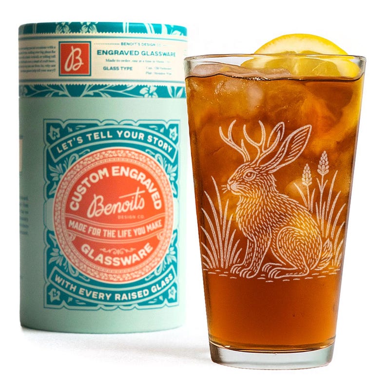 May include: A tall glass of iced tea with a lemon wedge, next to a cylindrical container. The glass is etched with a rabbit design. The container is light blue with the text "Custom Engraved Glassware".