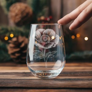 以下が含まれることがあります： 白いポピーの花がエッチングされた透明なガラス製のワイングラス。グラスは木製の表面の上に置かれています。