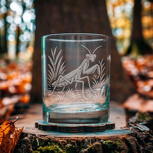 May include: A clear glass tumbler with an etched design of a praying mantis and tall grass. The glass sits on a wooden coaster, with a blurred forest background. The tumbler is approximately 3.5 inches tall.