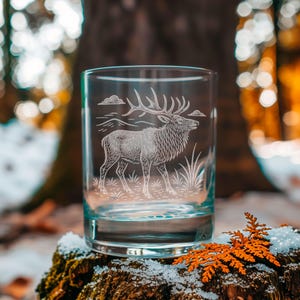 May include: A clear glass tumbler with an etched design of a stag, clouds, and grass. The glass is cylindrical and sits on a mossy surface with snow and orange leaves. The background is blurred, suggesting a forest setting.