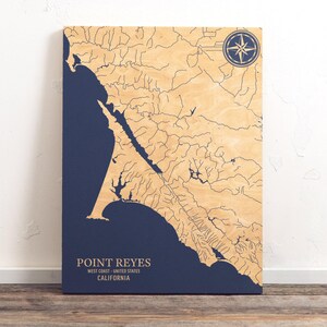 Pode incluir: Um mapa de madeira de Point Reyes, Califórnia, com uma rosa dos ventos no canto superior direito. O mapa está sobre um fundo azul e mostra a costa e os rios da área. O texto "POINT REYES" está impresso abaixo do mapa, juntamente com "WEST COAST - UNITED STATES" e "CALIFORNIA".