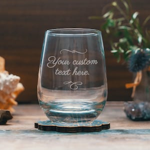 Puede incluir: Una copa de vino sin tallo de vidrio transparente con el texto "Your custom text here." grabado en el lateral. La copa está sobre un posavasos de madera con un diseño azul y blanco.