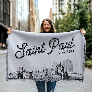 May include: A gray woven throw blanket with a black and white illustration of the Saint Paul, Minnesota skyline. The text "Saint Paul, Minnesota" is printed in black on the blanket.