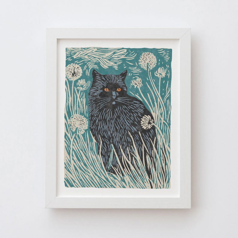 May include: A framed art print featuring a black cat with orange eyes, set amidst tall grass and white dandelions. The background is a teal color with a stylized sky. The artwork is in a white frame.