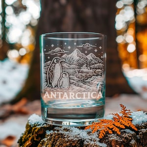 May include: A clear glass tumbler with a white etched design featuring two penguins, mountains, and a ship, with the word "ANTARCTICA" below. The glass is set on a mossy surface with snow and orange foliage.