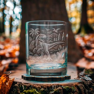 May include: A clear glass tumbler with a detailed etched design of a dinosaur, palm trees, mountains, and birds. The glass sits on a wooden coaster. The background features a blurred forest scene with fall foliage.