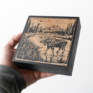 May include: A woodblock print of a moose standing in a lake with mountains in the background. The print is framed in black wood.