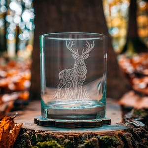 Può includere: Un bicchiere trasparente con un'immagine incisa di un cervo. Il cervo è in piedi sull'erba e il bicchiere è appoggiato su un sottobicchiere di legno. Lo sfondo è una scena sfocata di una foresta con foglie autunnali.