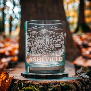 May include: A clear glass tumbler with a detailed white etched design featuring mountains, a guitar, a building, and a beer glass. The word "ASHEVILLE" is etched at the bottom. The glass sits on a wooden coaster, with a blurred forest background.
