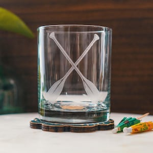 May include: A clear, cylindrical drinking glass with an etched design of two crossed oars. The glass sits on a decorative coaster with a floral pattern. Cocktail umbrellas are in the background.