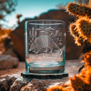 May include: A clear glass tumbler with an etched design of an armadillo, cactus, and desert landscape. The glass sits on a wooden coaster, with a blurred desert background and a cactus in the foreground.
