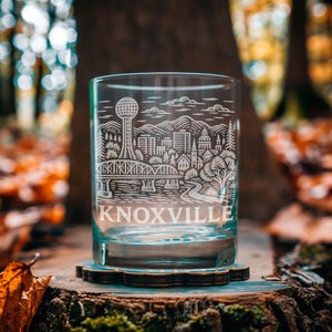 Puede incluir: Un vaso de vidrio transparente con un diseño grabado detallado del horizonte de Knoxville, que incluye la Sunsphere, puentes y edificios. La palabra "KNOXVILLE" está grabada en la parte inferior. El vaso está sobre un posavasos de madera.
