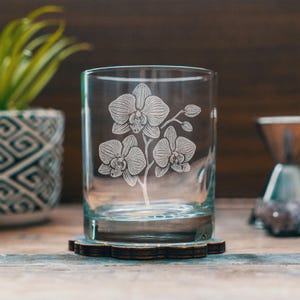 Op de afbeelding: Een helder glazen tumbler met een geëtst orchideeënontwerp. Het bloemmotief heeft drie orchideeën bloesems en een stengel. Het glas staat op een donkere houten onderzetter met een decoratieve rand. Een potplant en een metalen voorwerp staan op de achtergrond.