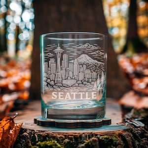 May include: A clear glass tumbler with a detailed etched design of the Seattle skyline, including the Space Needle and a mountain. The word "SEATTLE" is etched at the bottom. The glass sits on a wooden coaster, with a blurred forest background.