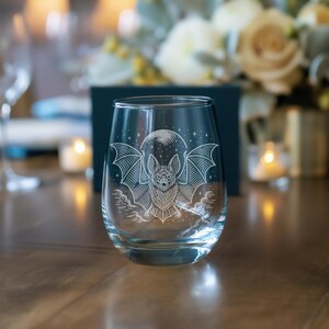 May include: A clear stemless wine glass featuring an etched design of a bat with outstretched wings, a full moon, and stars. The bat design is in white. The glass is on a wooden table with a blurred background of flowers and candles.