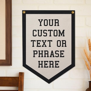 May include: A custom banner with a light beige background and black trim. The banner has the text "YOUR CUSTOM TEXT OR PHRASE HERE" in bold, black letters. Gold grommets are at the top corners for hanging.