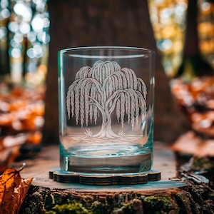 May include: A clear glass tumbler with a detailed etched design of a weeping willow tree. The glass sits on a wooden coaster, with a blurred forest background. The tree design is white, and the glass is filled with a clear liquid.