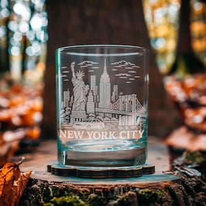 May include: A clear glass tumbler with a detailed etched design of the New York City skyline, including the Statue of Liberty, the Empire State Building, and the Brooklyn Bridge. The words "NEW YORK CITY" are etched below the skyline. The glass sits on a wooden coaster.
