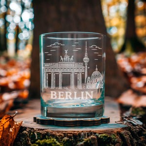 Peut inclure: Un verre transparent gravé avec des monuments emblématiques de Berlin, dont la porte de Brandebourg et la tour de télévision. Le mot "BERLIN" est gravé en dessous. Le verre est posé sur un dessous de verre en bois, avec un arrière-plan flou de forêt.