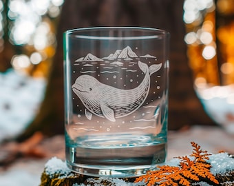 May include: A clear glass tumbler with a detailed etched design of a whale swimming in the ocean, with mountains in the background. The glass is cylindrical and sits on a mossy surface with snow and orange foliage.