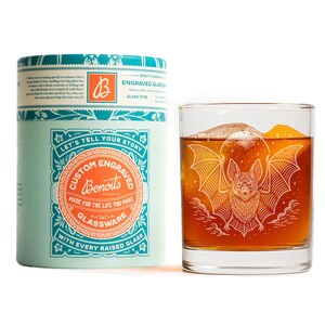 May include: A rocks glass with a bat engraving, containing a dark amber liquid and ice. Beside it is a teal and white cylindrical box. The box and glass are branded with the text "Custom Engraved Glassware".