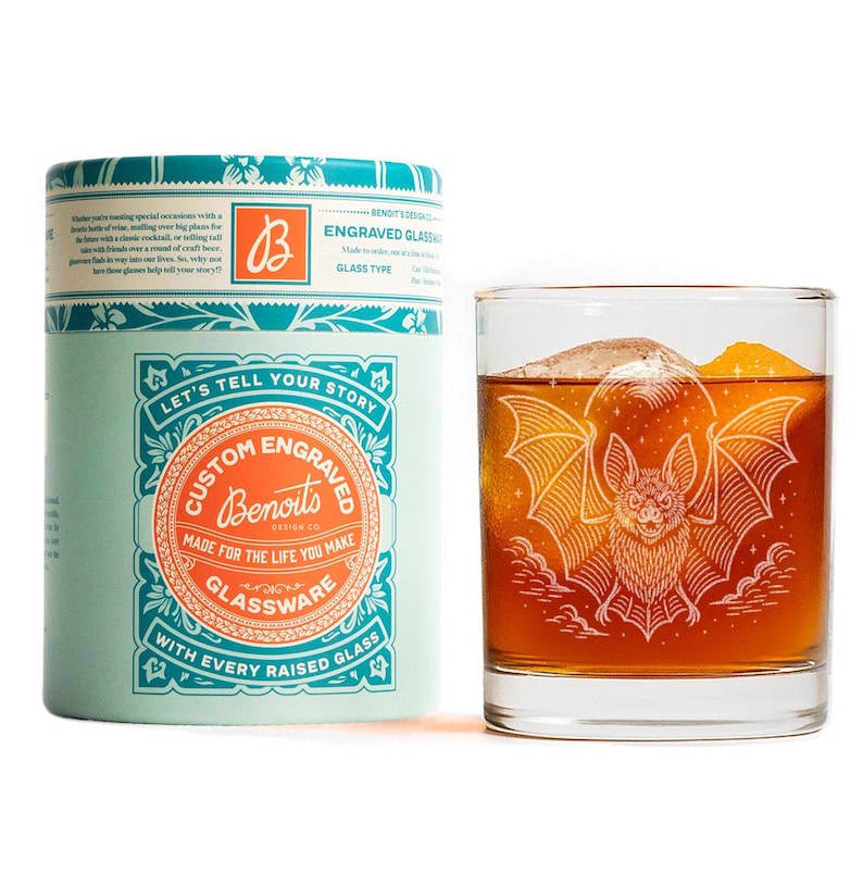 May include: A rocks glass with a bat engraving, containing a dark amber liquid and ice. Beside it is a teal and white cylindrical box. The box and glass are branded with the text "Custom Engraved Glassware".