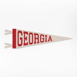 以下が含まれることがあります： 「GEORGIA」の文字が赤い太字で書かれた、クリーム色のペナント。ペナントの左側には赤い縁取りがあり、クリーム色のリボンが2本付いています。これはお土産や装飾品です。
