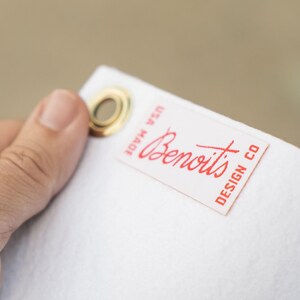 A hand holding a white tag with a red handwritten label that says "Benoits Design Co."